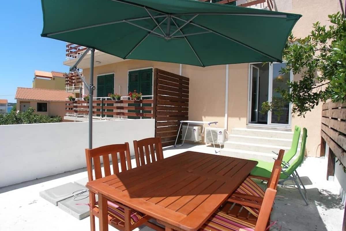 A wooden table and chairs under a green umbrella on a rooftop terrace with a view of buildings and a tree.