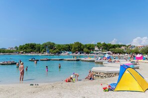 Egy tiszta, kékeskék vízű strand, ahol emberek úszkálnak, és a homokos partján egy sátor van felállítva.