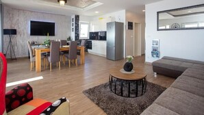 A modern living room with a sofa, a dining area with chairs, a kitchen with a refrigerator, and a flat-screen TV mounted on the wall. - Apartment Slavena (Okrug)