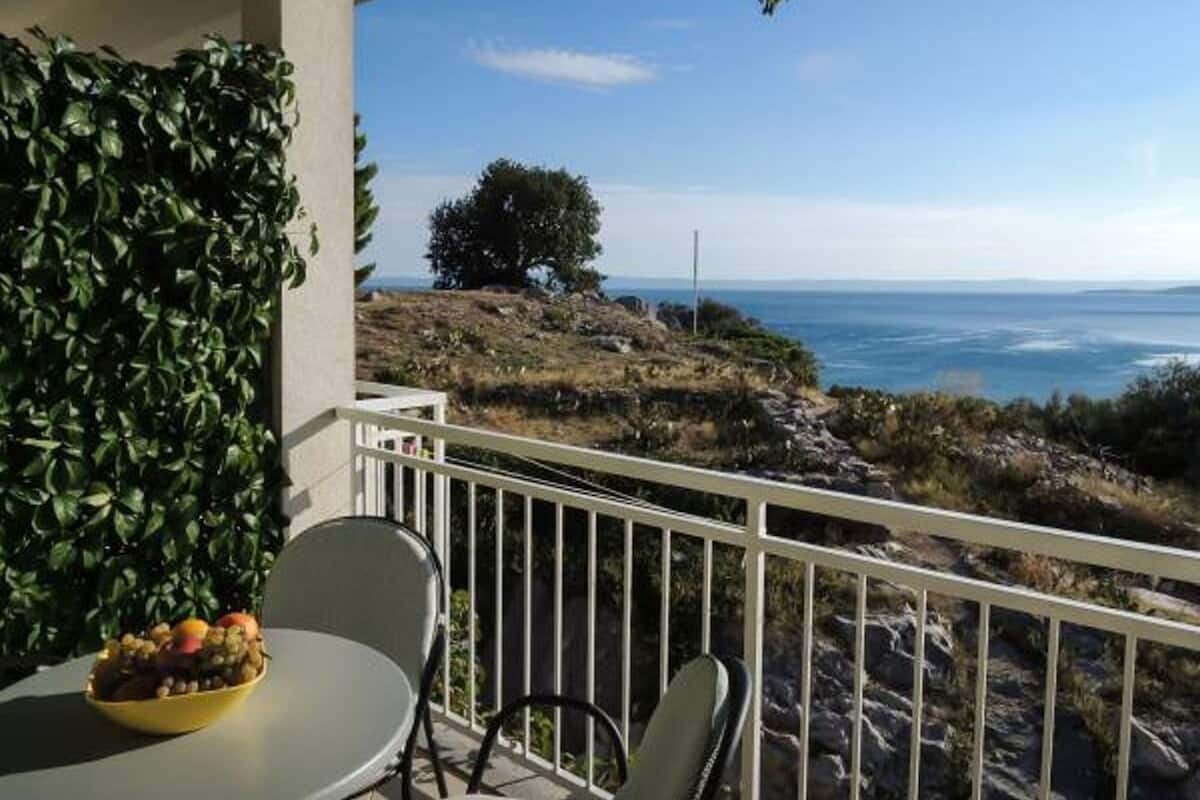 A balcony with a potted plant, a table with fruits, and a view of the ocean and rocky landscape.