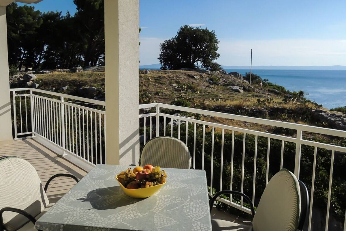 A balcony with a table and chairs, a bowl of fruit, and a scenic view of the sea and hills.