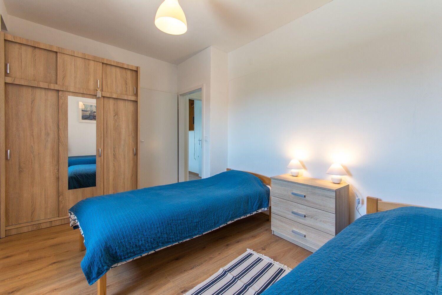 A bedroom with two beds, wooden wardrobe, and bedside lamps.