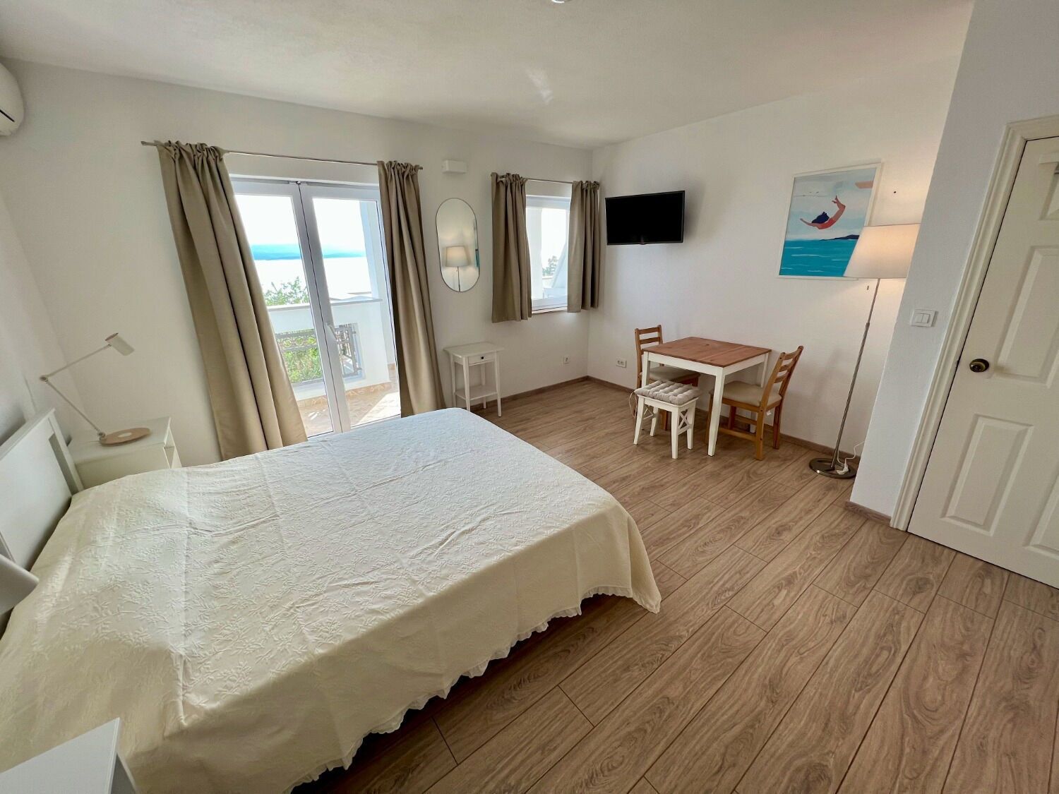 Un dormitorio con cama, escritorio, silla, televisor y balcón con cortinas.