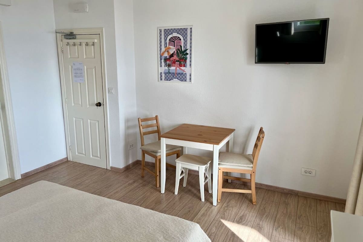 A room with a bed, a wooden table with two chairs, a TV mounted on the wall, and a door with a notice on it.