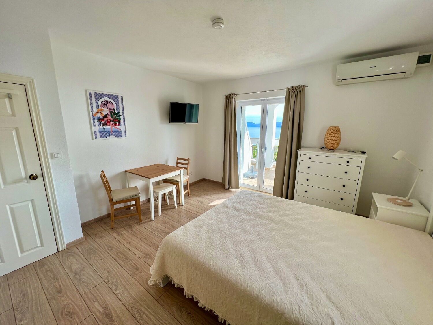 Een slaapkamer met een bed, een bureau met stoelen, een dressoir, een televisie en uitzicht op het groen.