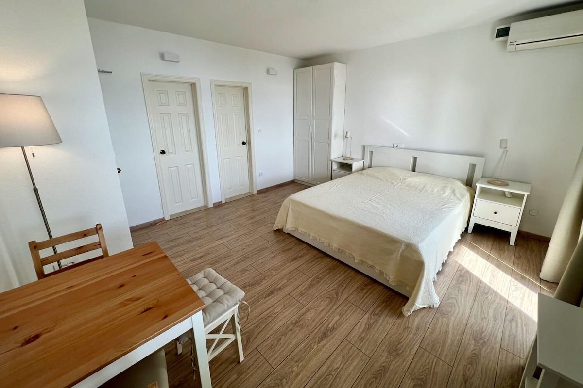 A bedroom with a bed, bedside tables, a desk, and a chair. There are two doors and an air conditioning unit on the wall.