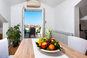 A balcony with a table set with fruit, a potted plant, and a view of buildings. - Apartments Marija (Rovinj)