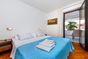 A hotel room with a bed, a nightstand, a wall-mounted lamp, a painting, and a sliding glass door to a balcony with a chair and a table. - Apartments Marija (Rovinj)
