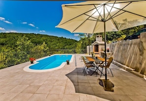 A swimming pool with a curved edge, surrounded by a tiled patio. There is a table and chairs under a large umbrella, and a small structure with a chimney. - House Brapa (Hrvace)