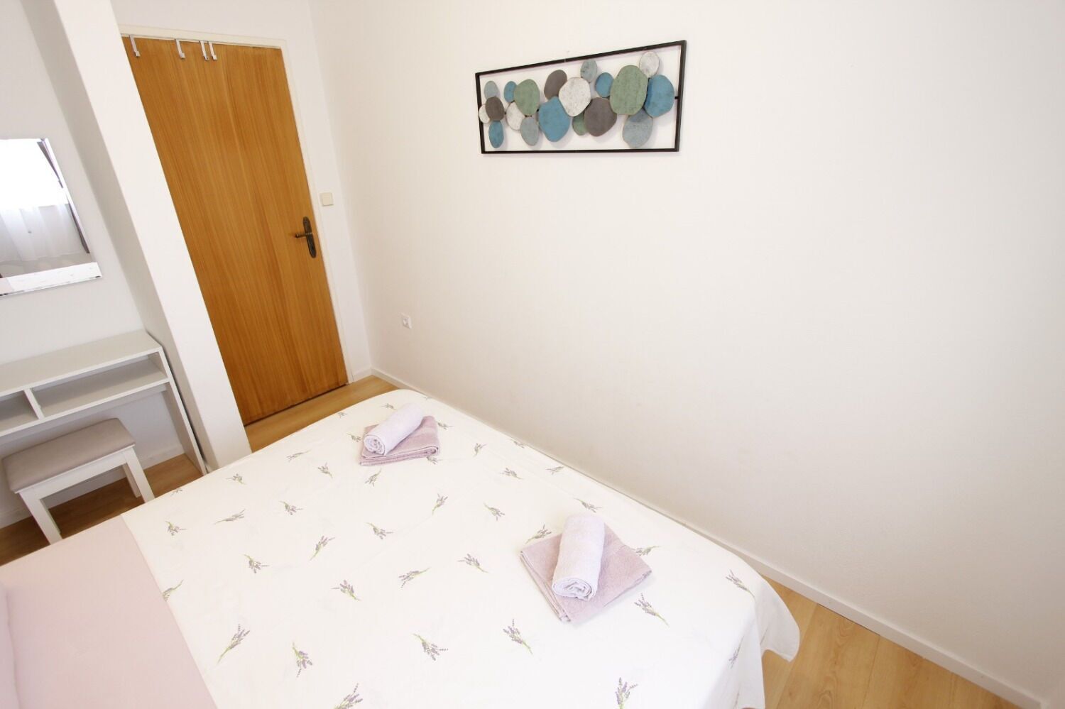 A bedroom with a bed, a wooden door, a white desk, and a wall-mounted artwork.