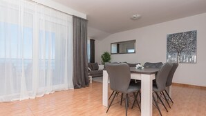 A modern dining area with a white table and grey chairs, a grey sofa, and a large window with sheer curtains. - Apartments Ivi (Podgora)