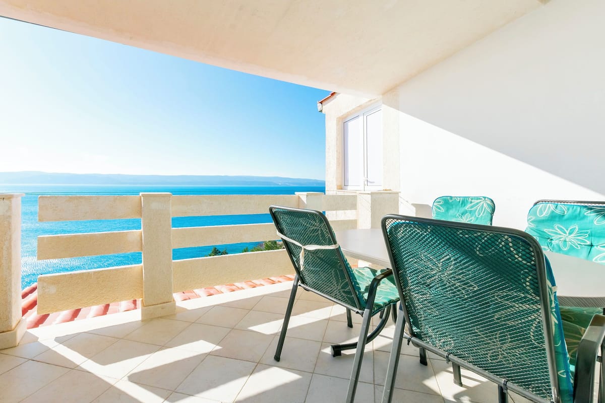 A balcony with a view of the sea, a table with chairs, and a white cabinet.