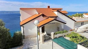 A two-story building with a red-tiled roof, labeled "APARTMANI," featuring a stone-paved entrance and a view of the sea. - Apartments Ivo (Omis)