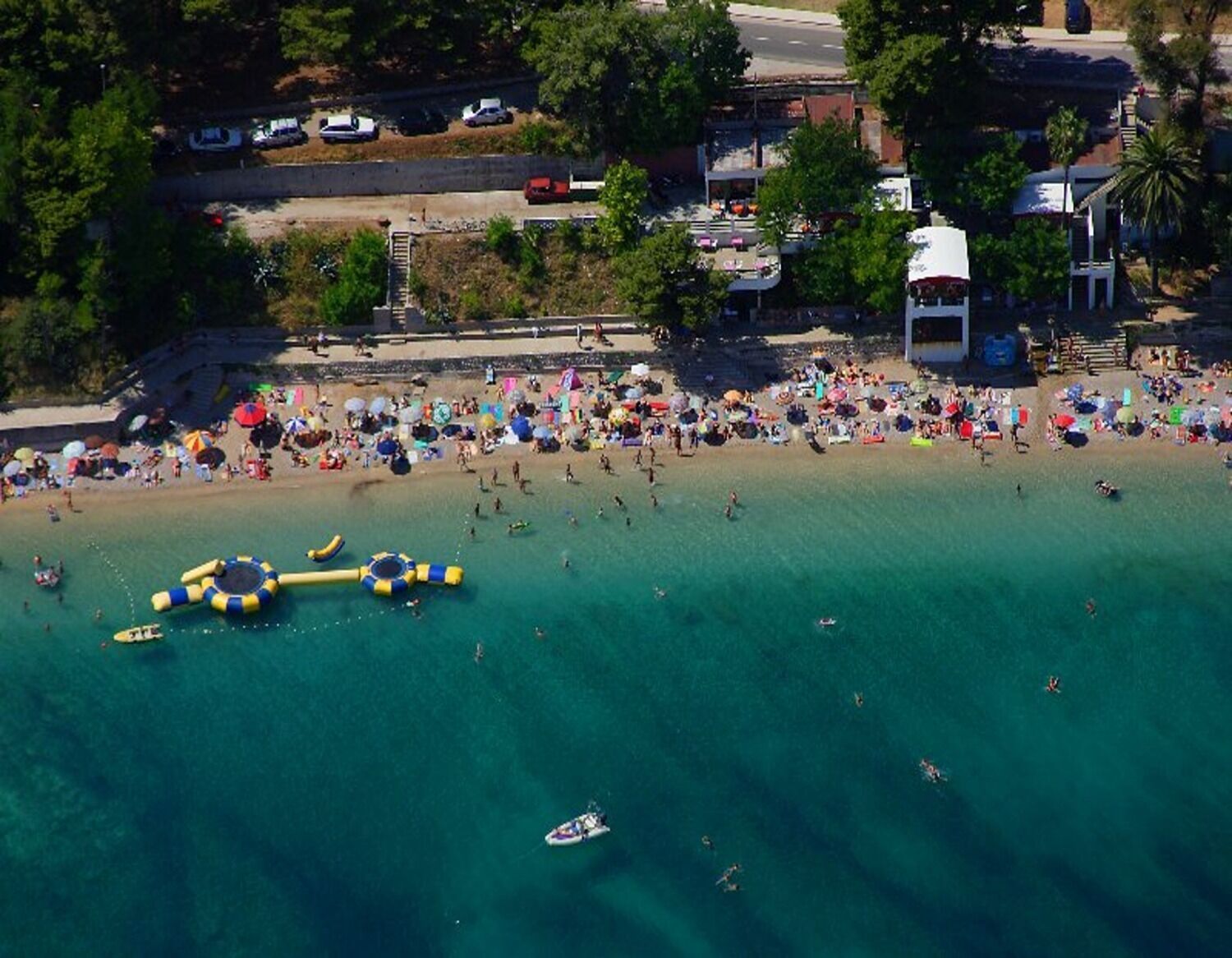 Luchtbeeld van een druk strand met helder turquoise water, opblaasbare constructies en een parkeerplaats op de achtergrond.
