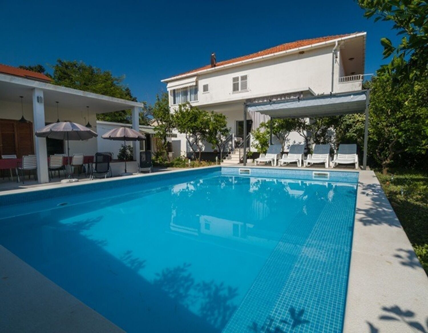 A swimming pool with lounge chairs and umbrellas, surrounded by a white building with a red-tiled roof.