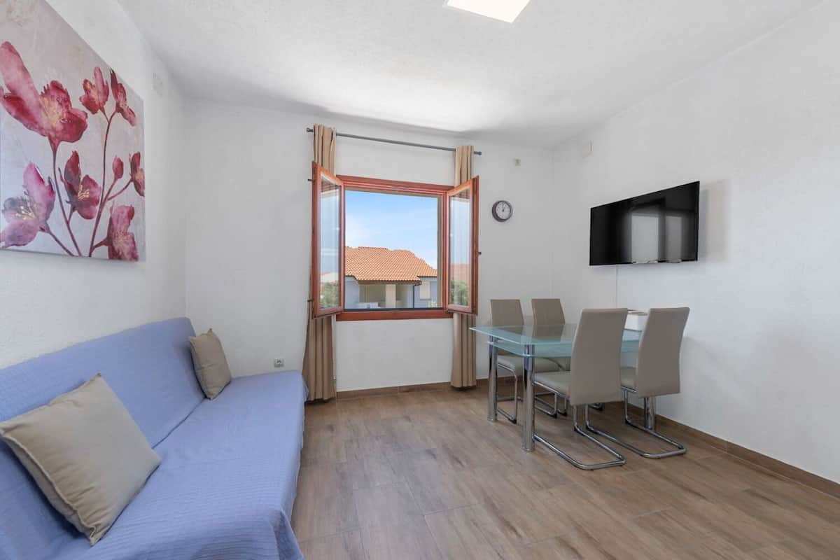 A room with a blue sofa, a glass dining table with chairs, a TV mounted on the wall, and a window with curtains.