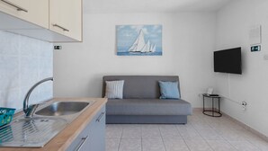 A compact kitchen with a sink, a small table, and a grey sofa with pillows. A painting of sailboats hangs on the wall, and a TV is mounted on the wall. - Apartment Uga2 (Kolan)