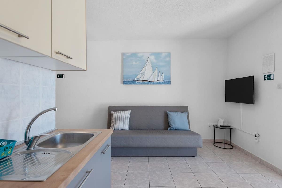 A compact kitchen with a sink, a small table, and a grey sofa with pillows. A painting of sailboats hangs on the wall, and a TV is mounted on the wall.