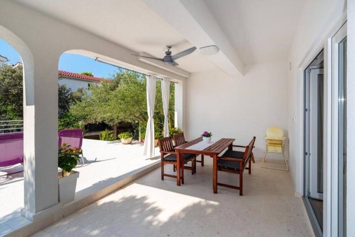 Un patio couvert avec une table et des chaises, un ventilateur de plafond et une vue sur la végétation environnante.