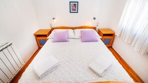 A double bed with a white textured bedspread, two purple pillows, and two white towels folded at the foot of the bed. There are two wooden nightstands with lamps, a framed picture on the wall, and a window with white curtains. - Apartments Bari (Kolan)