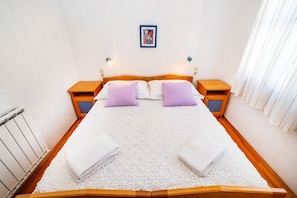 A double bed with a white textured bedspread, two purple pillows, and two white towels folded at the foot of the bed. There are two wooden nightstands with lamps, a framed picture on the wall, and a window with white curtains. - Apartments Bari (Kolan)