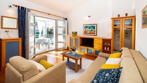 Ein geräumiges Wohnzimmer mit einer Couch, einem Couchtisch und einer TV-Konsole. Ein Balkon mit Tisch und Stühlen befindet sich außen.