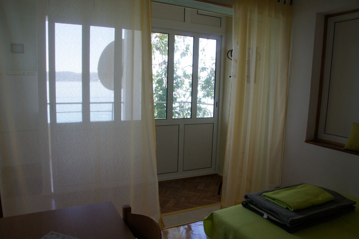 Ein Zimmer mit Tisch, Stuhl, Bett mit grüner Decke und einer Tür zu einem Außenbereich mit Blick auf Bäume und ein Gewässer.