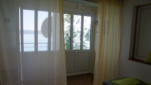 A room with a table, a chair, a bed with a green blanket, and a door leading to an outdoor area with a view of trees and a body of water. - Apartments Sonja (Podgora)