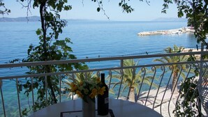 A balcony with a table, a bottle, and a vase of flowers overlooking a coastal view with clear blue water and distant land. - Apartments Sonja (Podgora)