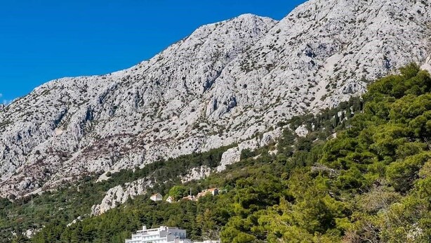 A coastal area with a clear turquoise sea, a sandy beach, and rocky cliffs. There are buildings with balconies overlooking the sea. - Apartments Sonja (Podgora)