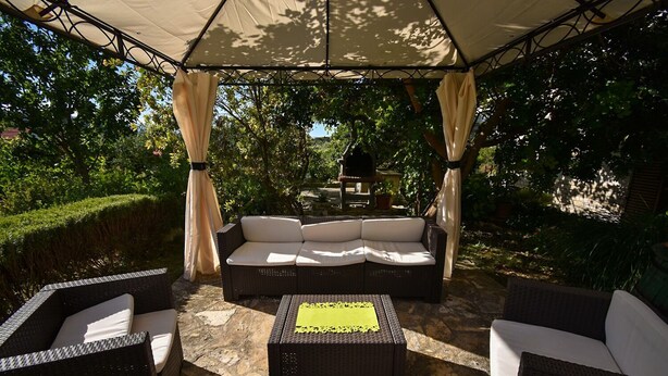 An outdoor patio with a covered seating area, stone flooring, and lush greenery in the background. - Apartments and Rooms Frane (Rab)