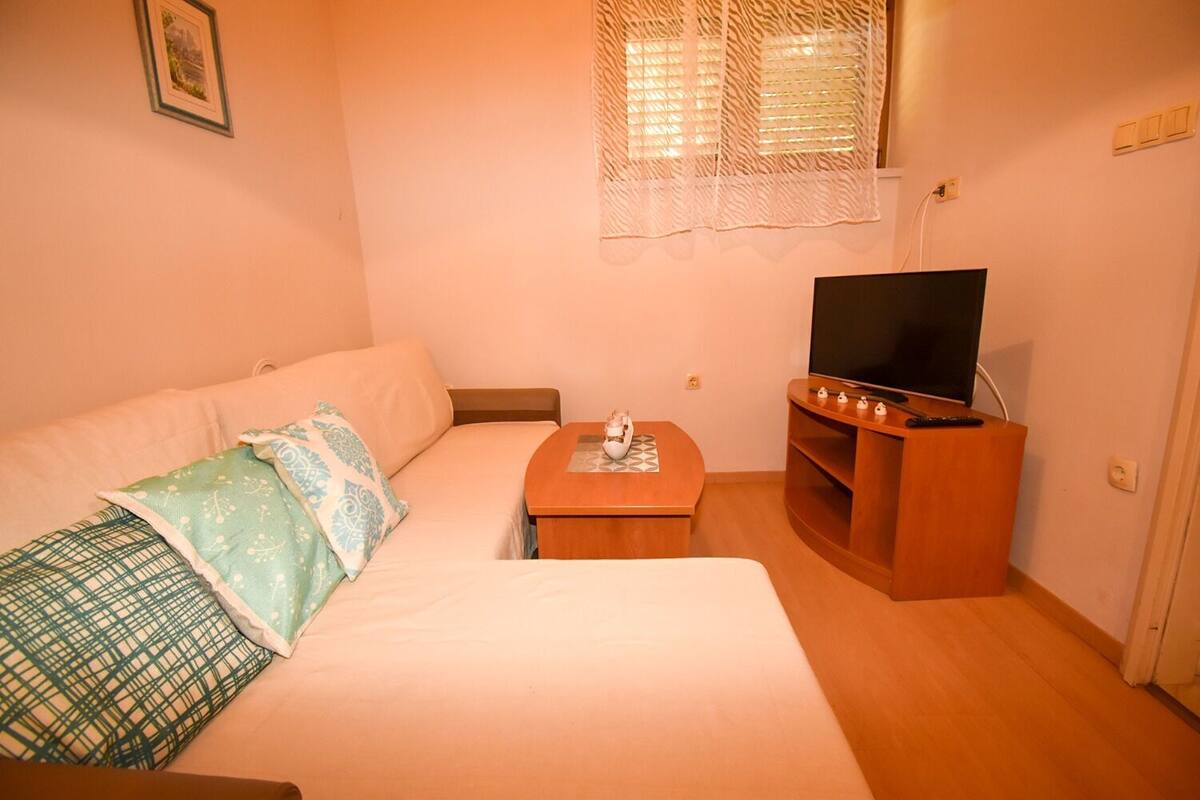 A small, cozy room with a bed, a TV on a wooden stand, and a window with blinds.