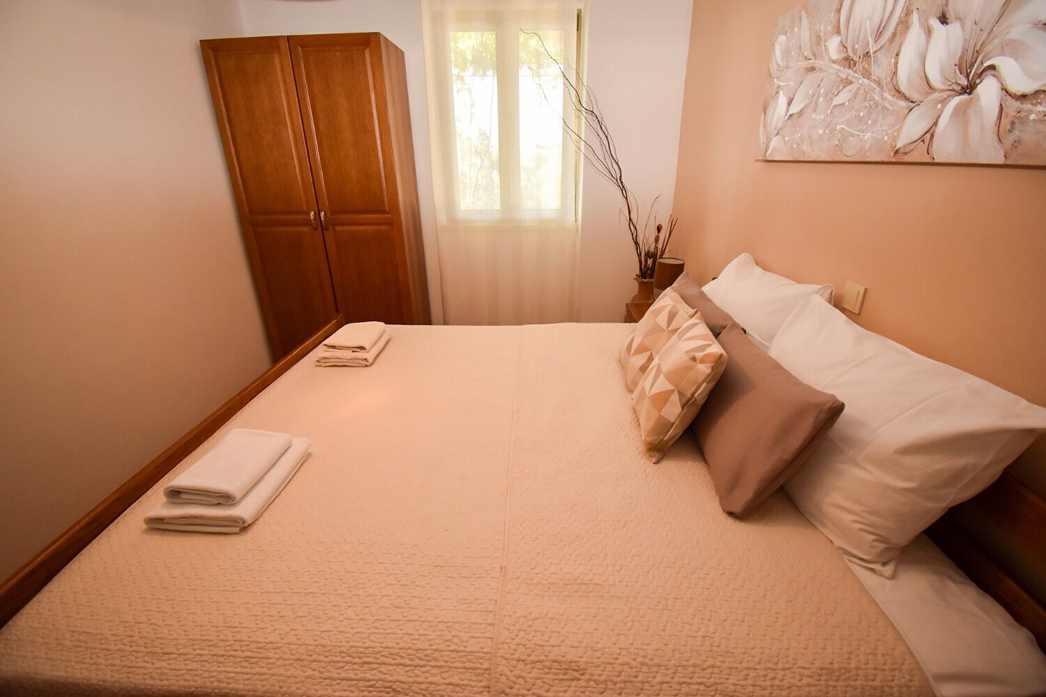 A neatly made bed with a beige bedspread and pillows, a wooden wardrobe, and a window with natural light.