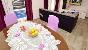 Un área de comedor con una mesa redonda, un cuenco de fruta y un mantel de encaje. Se ve un sofá y un televisor al fondo.