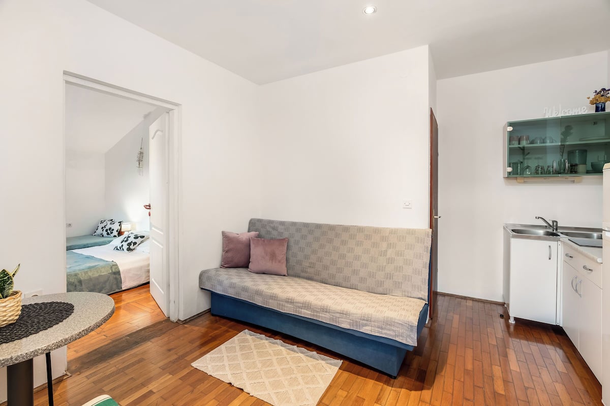 Ett kompakt bostadsutrymme med en soffa, ett litet bord, en köksdel och en synlig dörr som leder till ett annat rum.