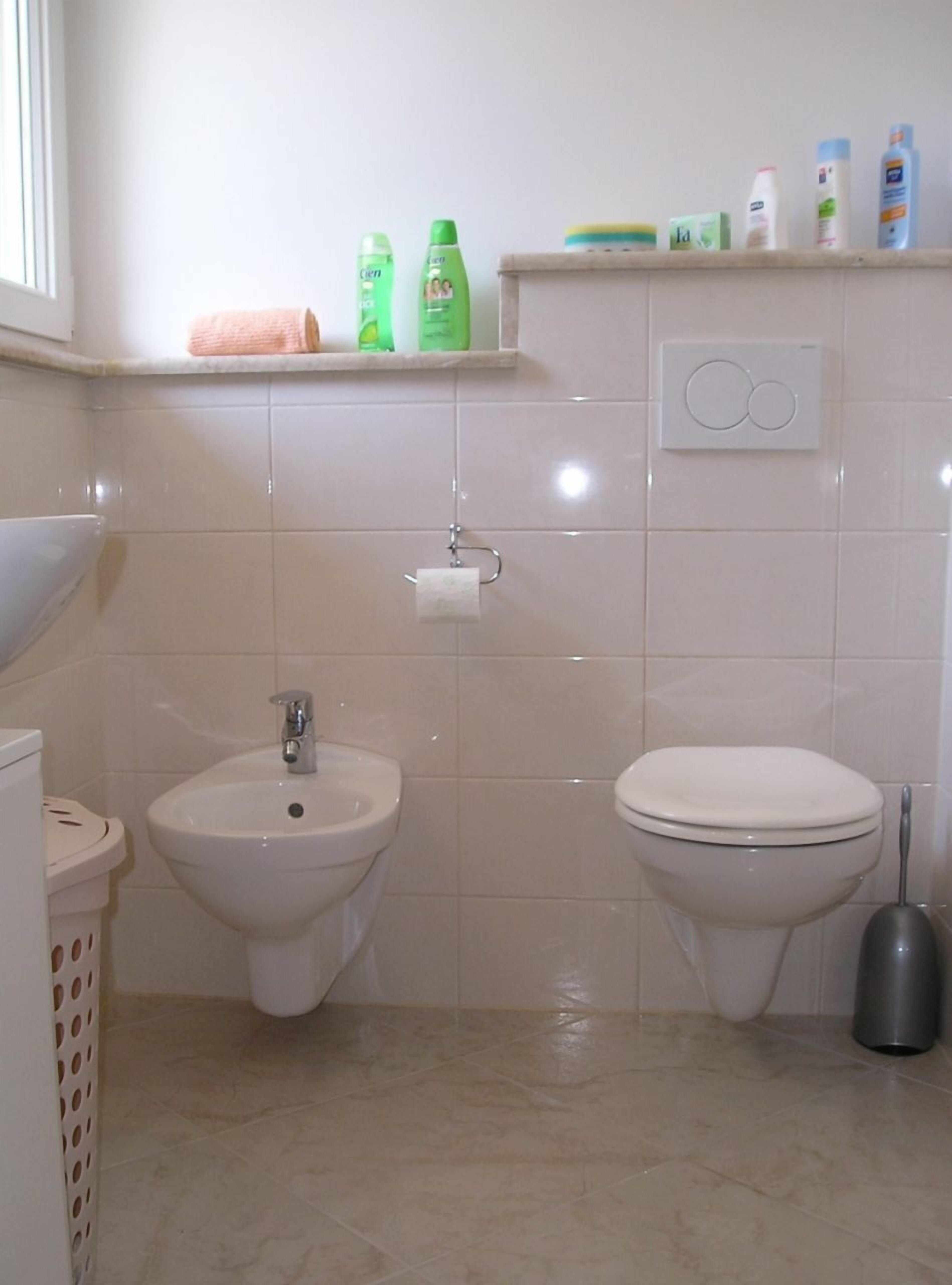 A bathroom with a bidet, toilet, sink, and a shelf with toiletries.