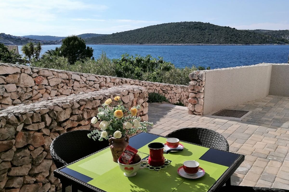 A table with a vase of flowers, a cup of coffee, and a teapot on a patio overlooking a body of water and hills.