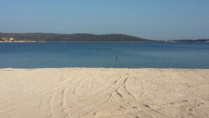 Ein sandiger Strand mit Fußspuren, die zu ruhigem, blauem Wasser führen, ein klarer Himmel und eine ferne Hügelkette.