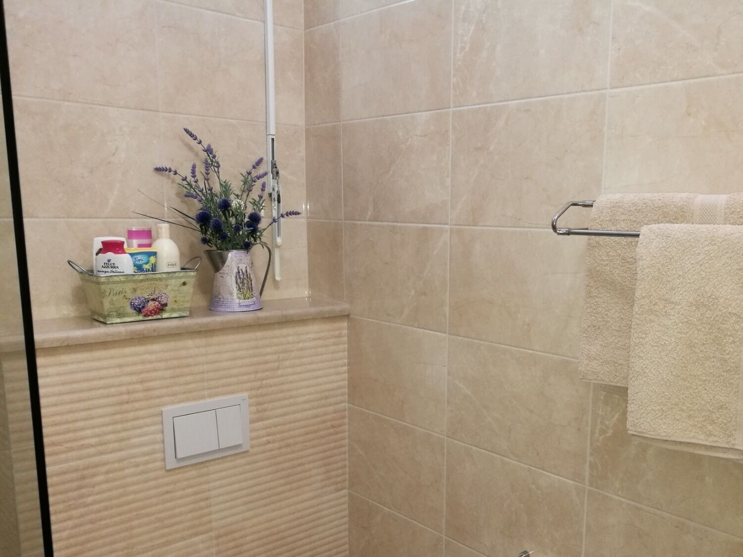 Un baño con baldosas color crema, ducha, toallero y estante con artículos de aseo y un florero.