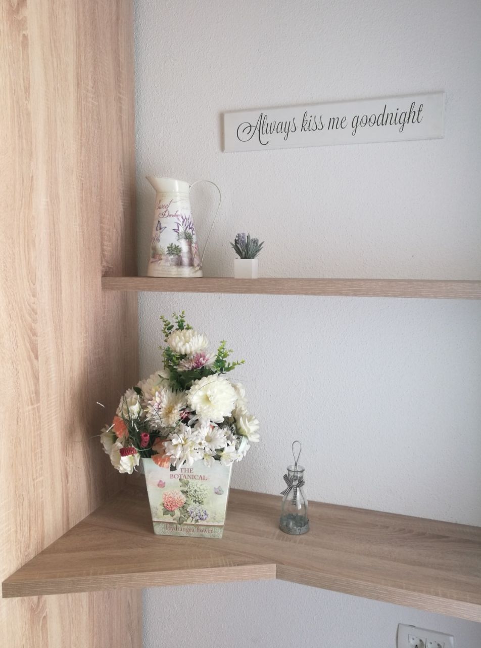En hylla i trä med en dekorativ kanna, en liten krukväxt, en blomsteruppsättning och en skylt med texten: ”Ge mig alltid en godnattskyss”.