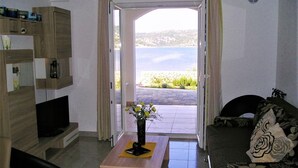 Ein Wohnzimmer mit einer Couch, einem Couchtisch mit einer Vase und einem Fernseher. Man sieht auf einen See oder ein anderes Gewässer und viel Grün.