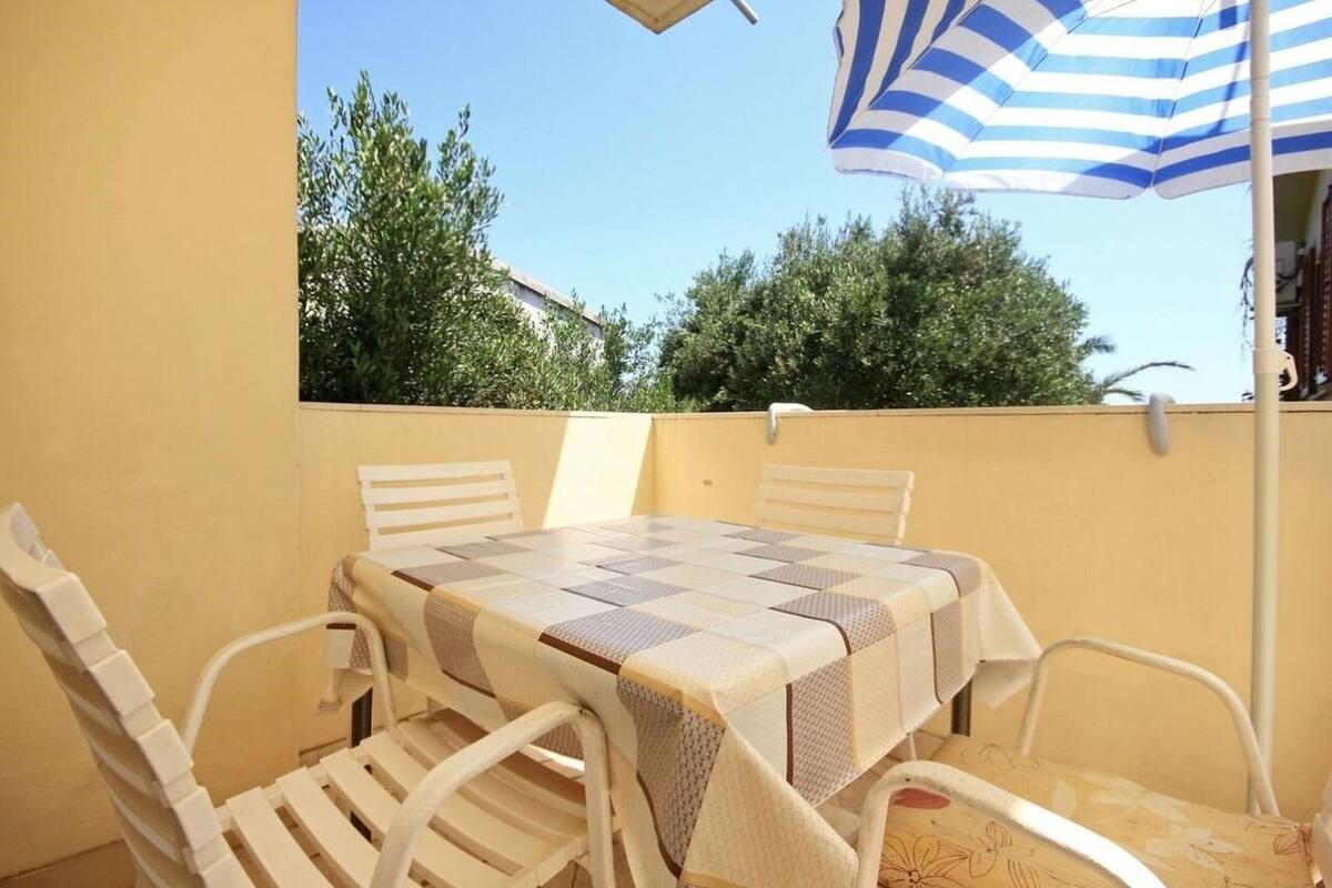 A table with a checkered tablecloth, surrounded by white chairs, under a striped umbrella.
