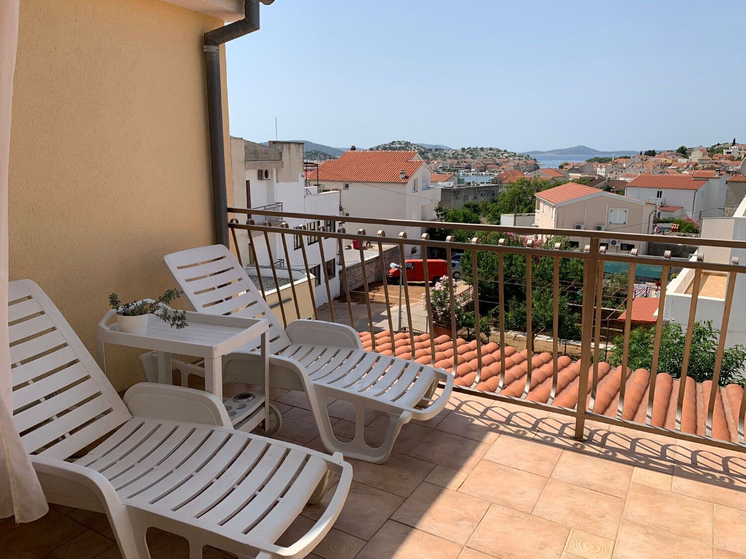 Een balkon met witte plastic stoelen en een kleine tafel, met uitzicht op een woongebied met terracotta daken en een verre zee.