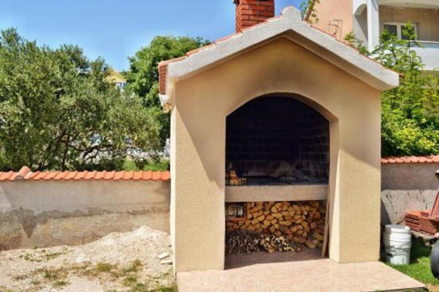 Ein kleiner, gemauerter Außenofen mit Schornstein, umgeben von einer niedrigen Mauer und einem Holzstoß.
