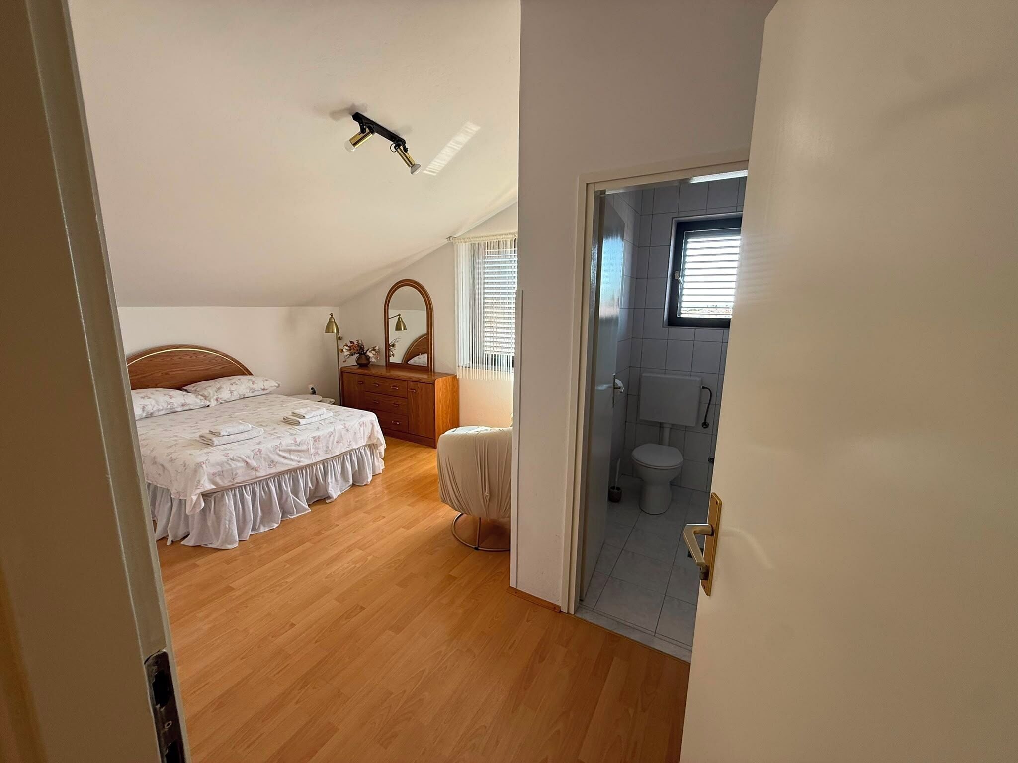 A bedroom with a wooden floor, a bed with a floral bedspread, a dresser, a mirror, and a ceiling light fixture.