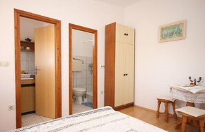Ein Schlafzimmer mit Bett, einem Holzschrank, einem kleinen Tisch mit Stuhl und einem durchgegangenen Türrahmen sichtbaren Badezimmer.