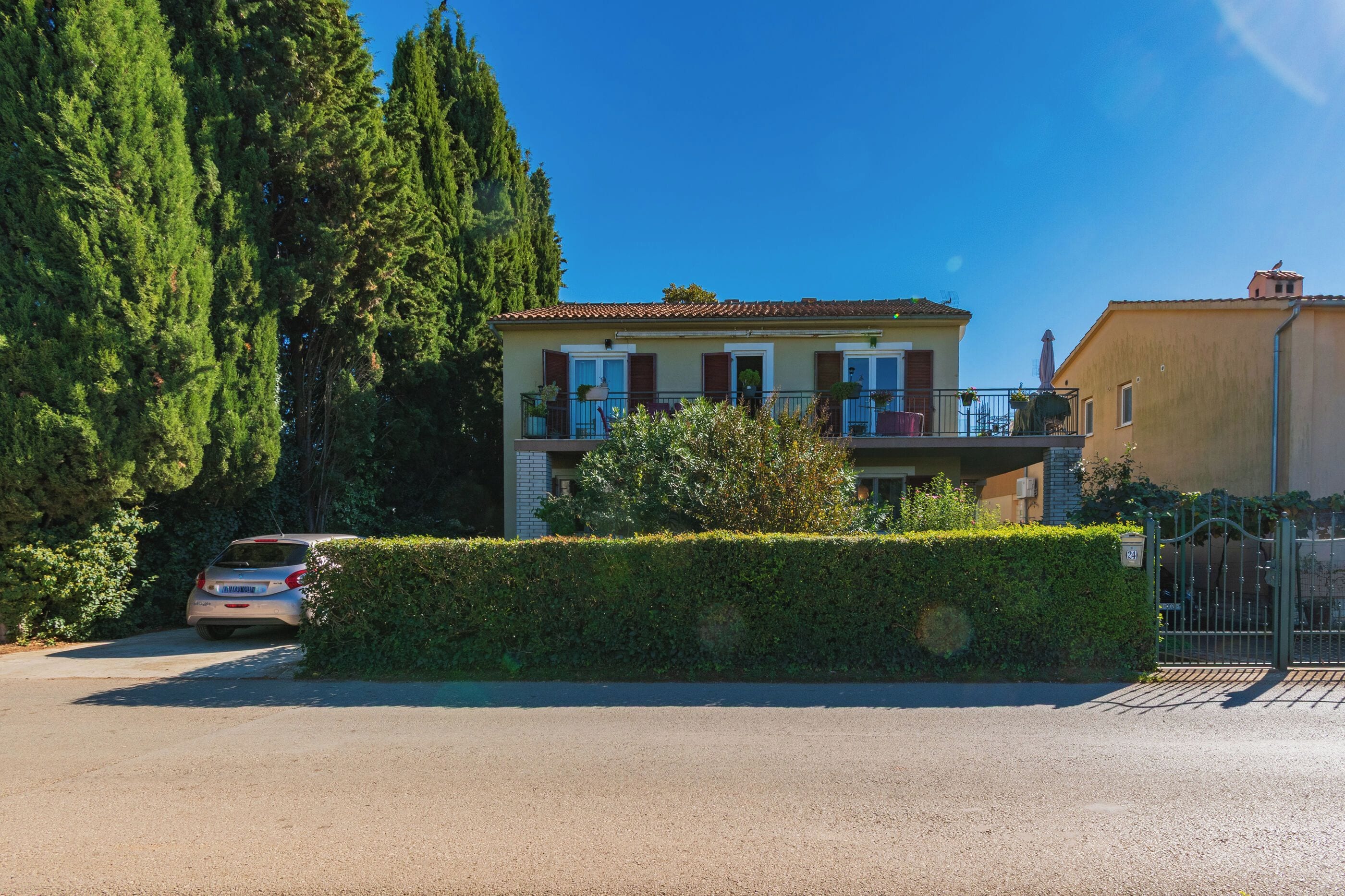 A two-story residential building with a balcony, a car parked in front, and a hedge in the foreground.