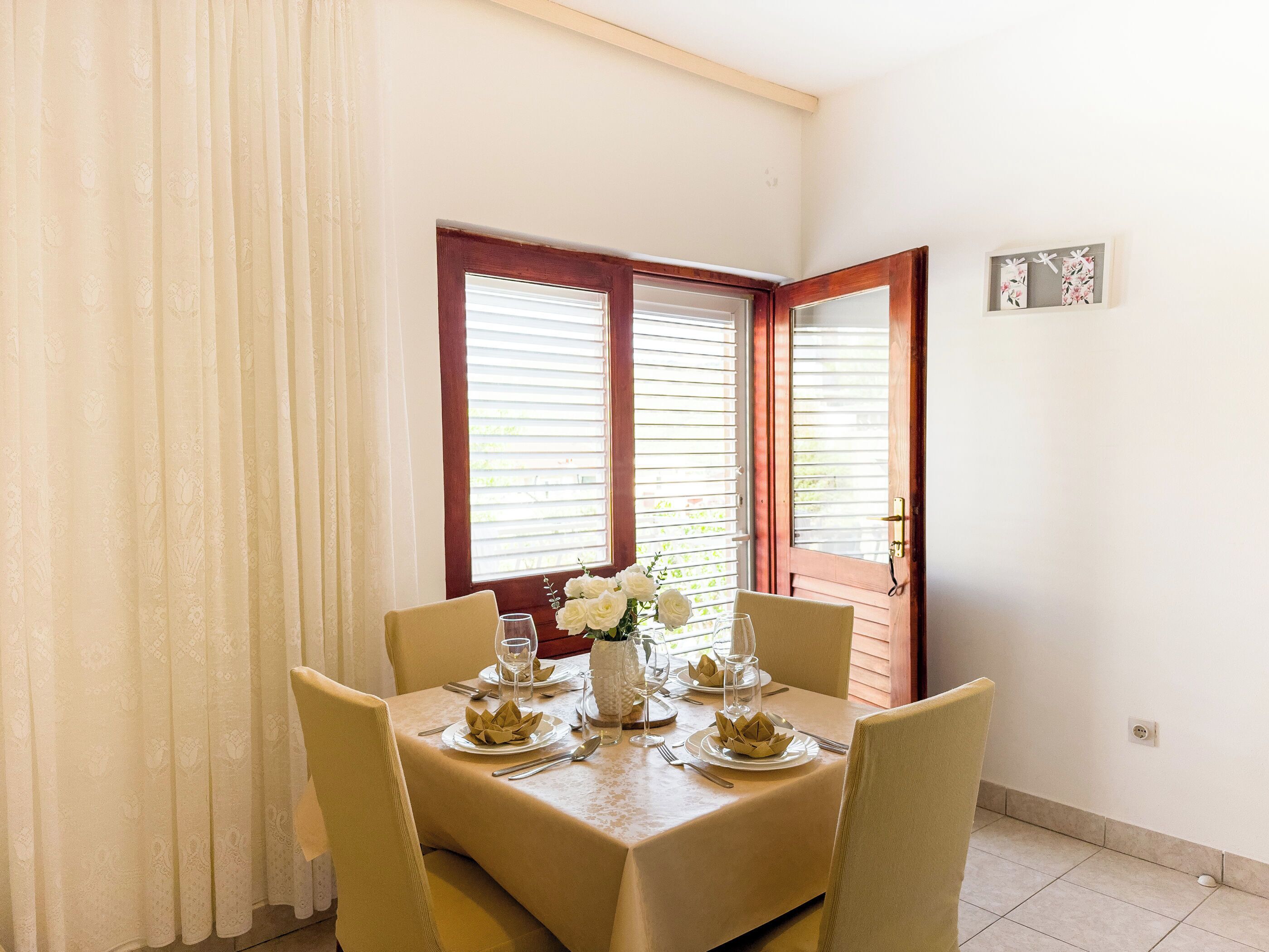 Un coin repas avec une table dressée pour quatre personnes, deux fenêtres équipées de stores et une porte avec une poignée.