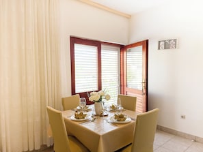 A dining area with a table set for four, two windows with blinds, a door, and a wall-mounted light switch. - Apartments Josi (Sibenik)
