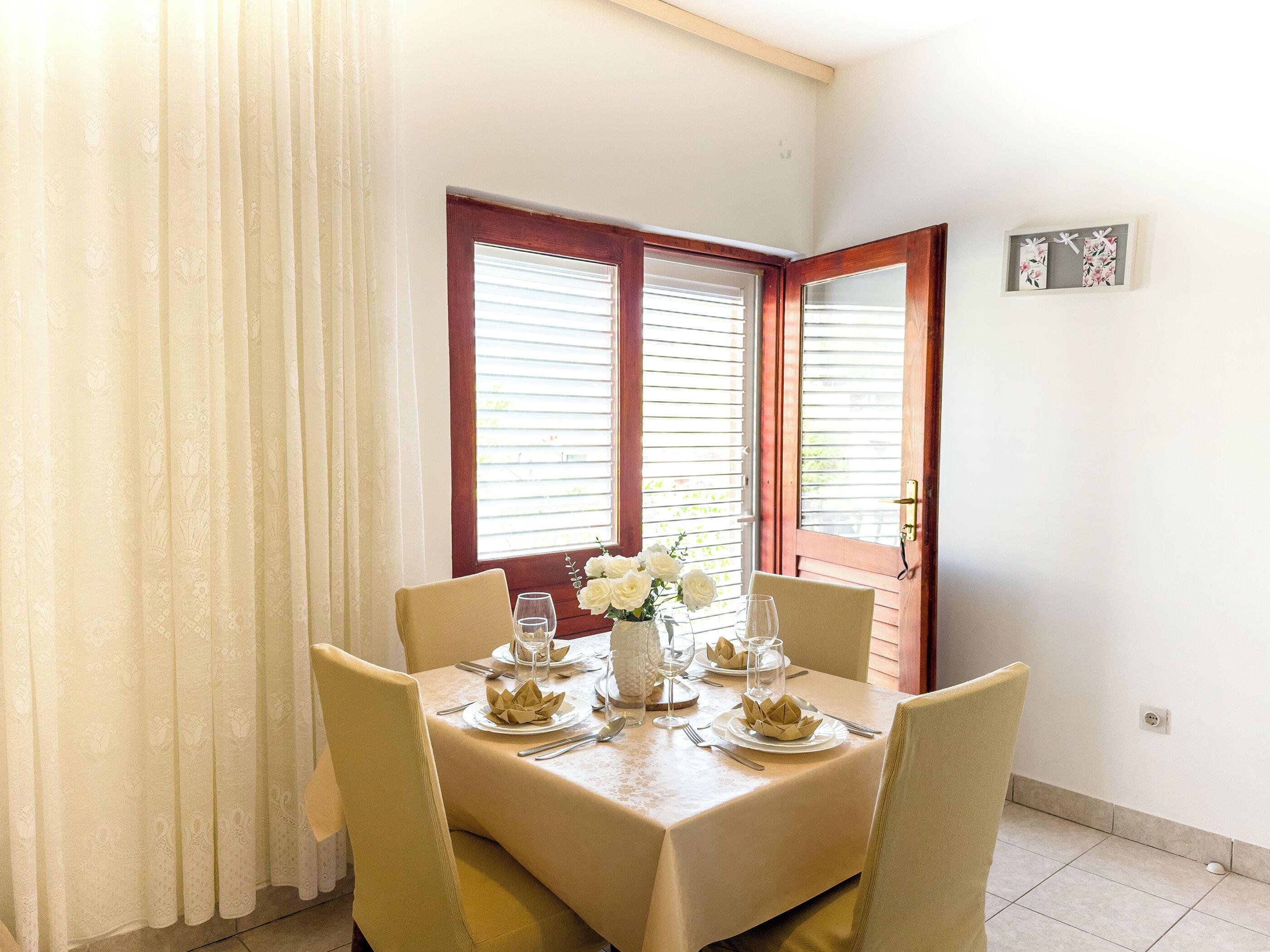 Un comedor con una mesa dispuesta para cuatro, un florero con flores blancas y una ventana con persianas.